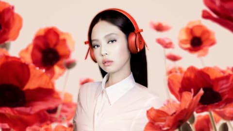 A professional promotional photograph featuring K-pop artist Jennie Kim positioned in the center, looking directly at the camera with a neutral expression. She has straight, dark hair, subtle makeup, and is wearing bright red Beats Solo 4 on-ear wireless headphones that feature small, decorative red ribbons tied on each side of the headband. She is dressed in a pale pink, semi-sheer, collared button-up shirt with ruched detailing across the front. The background is a soft, light beige color, heavily framed in both the foreground and midground by vibrant, slightly out-of-focus red poppy flowers with green stems, creating a layered, artistic floral depth. Jennie Kim stars in a vibrant promotional campaign wearing red Beats Solo 4 headphones surrounded by red poppies.