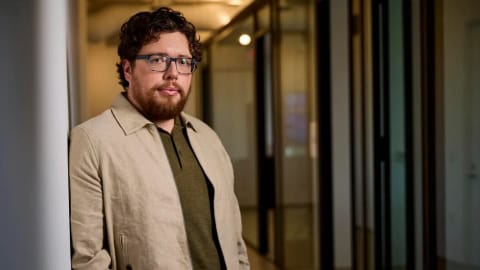Michael Sarnoski leaning against a stark white structural pillar on the left side of the frame. He is positioned inside a modern office or studio setting, with a long, brightly lit hallway trailing off into a soft-focus background behind him. He wears clear, square-rimmed eyeglasses, an olive-green polo shirt, and a light beige unbuttoned zip jacket. Sarnoski has wavy dark brown hair, a full, well-trimmed brown beard, and a calm, direct expression as he looks directly into the camera. The corridor behind him is framed with glass-paned doors and dark metal trim, catching warm ambient light that creates a clean, architectural depth.
