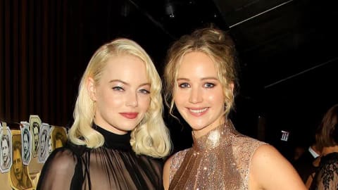 An eye-level indoor full shot features actresses Emma Stone and Jennifer Lawrence standing next to each other at the NYC premiere after-party for the movie Mother!. On the left, a smiling Emma Stone sports platinum blonde locks and wears a Valentino ensemble featuring a tiny black bandeau with a high-necked, sheer long-sleeved shirt over a floor-length black skirt. Her face is turned slightly towards the right as she stands near Jennifer Lawrence. On the right, Jennifer Lawrence is smiling while posing with one hand on her hip. She wears a sparkly, sleeveless, form-fitting gold and metallic-striped Ralph & Russo dress with high slits and fringe detailing at the hem. Her blonde hair is styled in a loose updo. In the background on the far right, another woman carrying a large reddish-brown leather tote bag stands near a table with white candles. The setting appears to be a dimly lit venue with wood paneling visible on the left wall.