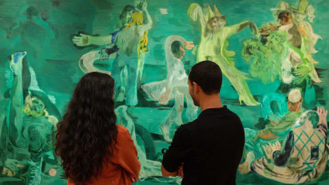 A wide gallery shot captured inside The Courtauld Gallery shows two gallery visitors—a woman with long, dark curly hair wearing an orange jacket on the left and a man in a black long-sleeved shirt on the right—standing with their backs to the camera as they closely study a massive figurative painting. The artwork, titled Ghost Ball (2023) by celebrated artist Salman Toor, dominates the frame with its signature, surreal palette of deep emerald and absinthe greens. Across the vast canvas, a series of loose, fluid, and elongated figures are depicted in various expressive, theatrical poses that suggest a dreamlike nocturnal gathering or party. The figures—some wearing whimsical caps or costumed elements, others lounging or dancing—inhabit a stylized, safe nocturnal environment that evokes themes of community, intimacy, and queer South Asian experiences. The bright gallery lighting illuminates the clean white wall surrounding the massive canvas, casting a subtle warmth on the shoulders of the spectators engrossed in the scene.