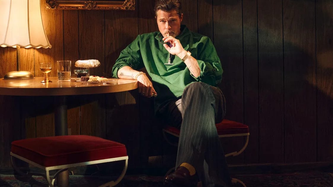 A visually rich, cinematic photograph captures actor Brad Pitt seated in a retro, wood-paneled room with a moody, warm ambiance. He wears a vibrant green pullover, pinstriped trousers, and brown loafers while holding a cigar near his mouth. He sits cross-legged next to a small wooden table illuminated by a scalloped-edge lamp, displaying glasses and pastries. Behind him, an ornate gold-framed portrait of a blonde woman hangs on the dark wood wall, and a large matching mirror reflects a glowing chandelier. The setting features a classic Persian rug on the floor and a plush red stool in the foreground, evoking a nostalgic, mid-century lounge aesthetic.