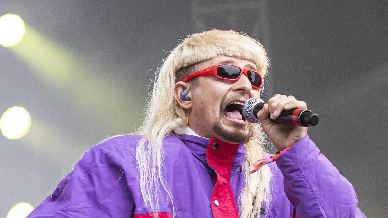 An eye-level medium close-up shows Oliver Tree performing live on an outdoor stage filled with a hazy atmosphere. The artist is singing passionately into a black microphone held in his right hand while facing slightly to the right. He features a distinctive blonde mullet with blunt bangs, a goatee, and bright red sunglasses. He wears a vibrant purple and red color-blocked windbreaker jacket, while blurred bright stage lights glow softly in the background.