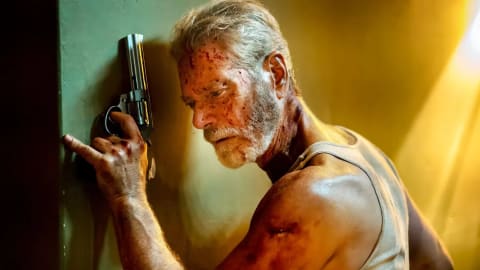Stephen Lang stars as the formidable Norman Nordstrom, a blind military veteran navigating intense combat in a dimly lit, sun-streaked interior hallway. In this high-contrast cinematic shot from the horror-thriller film Don't Breathe 2, Nordstrom leans heavily against a dark wall, his expression a mix of fierce concentration and physical exhaustion. His face, arms, and white tank top are heavily stained with blood and grime, highlighting the brutality of the conflict. He holds a large, silver revolver upward with a steady grip, his finger resting near the trigger as he listens intently for approaching threats. A warm, harsh light streams from the right background, cutting through the shadows and illuminating the dust in the air, emphasizing the gritty and suspenseful atmosphere of his isolated environment.