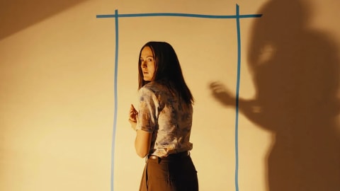 Renate Reinsve stars as Dr. Mary Kline in a suspenseful, dimly lit sequence from A24's psychological sci-fi horror feature Backrooms, looking back over her shoulder with an expression of intense vulnerability and fear. Clad in a patterned, short-sleeved short button-up shirt and high-waisted brown trousers, she stands frozen against a warm, yellow-toned wall bisected by a crude doorway outlined in bold blue tape. Looming aggressively over the right side of the frame is a stark, distorted human shadow cast onto the wall surface, its hand raised in a tense or demanding gesture. The sharp cinematic lighting emphasizes the depth of the shadow and highlights the actress's panicked profile, capturing the exact moment the film's domestic reality fractures into a surreal, multi-dimensional nightmare.