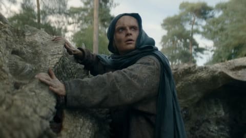 A heavily scarred, wide-eyed Aegon II Targaryen, portrayed by Tom Glynn-Carney, is shown hiding in disguise during his escape from King's Landing in House of the Dragon. He is wearing a dark, hooded teal-grey cloak over a patterned brown medieval robe. His face bears severe burn scars and disfigurement from the Battle of Rook's Rest. He grips the textured surface of a large, fallen moss-covered log or stone formation with both hands as he crouches low in a rocky, trench-like depression. His expression is one of sudden alarm and vulnerability as his eyes look upward and out of frame, scanning for potential threats. The background features a soft-focus forest with thin pine trees against a pale, overcast sky, establishing a bleak and tense outdoor environment. 