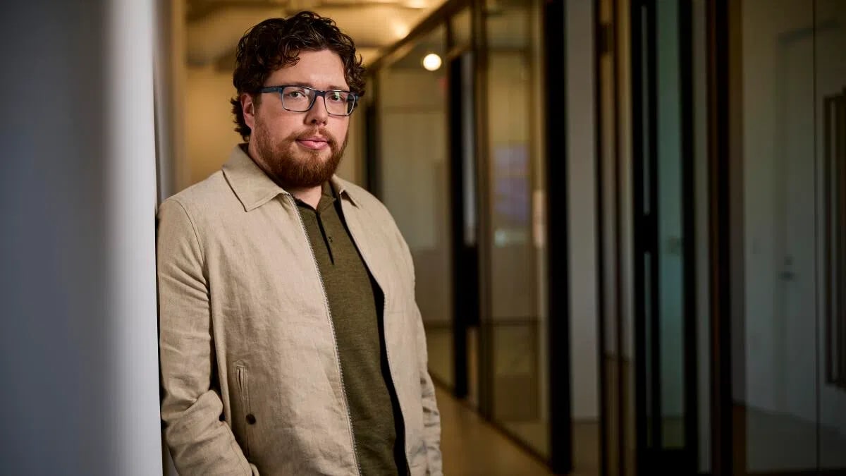 Michael Sarnoski leaning against a stark white structural pillar on the left side of the frame. He is positioned inside a modern office or studio setting, with a long, brightly lit hallway trailing off into a soft-focus background behind him. He wears clear, square-rimmed eyeglasses, an olive-green polo shirt, and a light beige unbuttoned zip jacket. Sarnoski has wavy dark brown hair, a full, well-trimmed brown beard, and a calm, direct expression as he looks directly into the camera. The corridor behind him is framed with glass-paned doors and dark metal trim, catching warm ambient light that creates a clean, architectural depth.