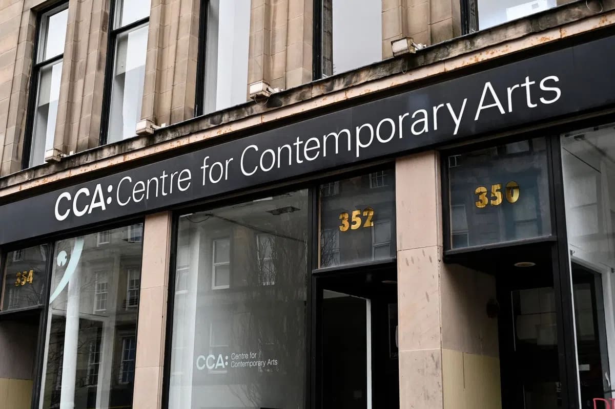 A black sign featuring the words "CCA: Centre for Contemporary Arts" in bold white lettering hangs prominently above the entrance of a stone building with large, dark-framed display windows and glass doors reflecting surrounding architectural details. A horizontal, eye-level shot captures the exterior entrance of the Centre for Contemporary Arts (CCA) building, focusing on a long, sleek black signboard with the institution's name written in crisp, clean white sans-serif font. Below the sign, large transparent glass windows and glass entrance doors are set within a pale stone facade, reflecting the outlines of historical buildings across the street, while gold numbers "352" and "350" are visible on the glass panels above the doorways.
