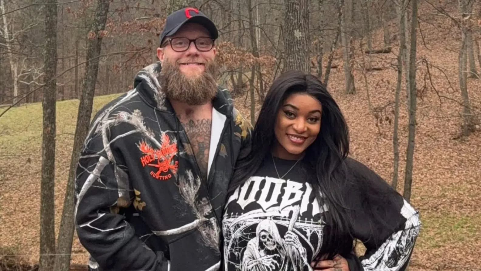 A medium outdoor shot features the social media influencer couple Dalton Memel (known online as "Dalton Ain't Worried") and his partner, Ntsako "Sako" Mathivha, standing close together in front of a wooded backdrop with bare trees and fallen brown leaves covering a sloped hillside. Dalton, standing on the left, is a Caucasian man with a thick, well-groomed brown beard, wearing glasses, a black baseball cap with a red logo, and a black zip-up hoodie featuring a white-and-grey tree branch camouflage pattern over a graphic design that reads "HOLLY RIVER HUNTING CLUB". Sako, standing on the right, is a Black woman with long, dark, wavy hair parted down the middle, wearing a dark silver necklace and a black long-sleeved graphic sweatshirt featuring a skeleton holding a scythe under an arched structure with gothic typography above it. They are both looking directly at the camera with gentle, confident smiles, posed comfortably together against the natural rural background. 