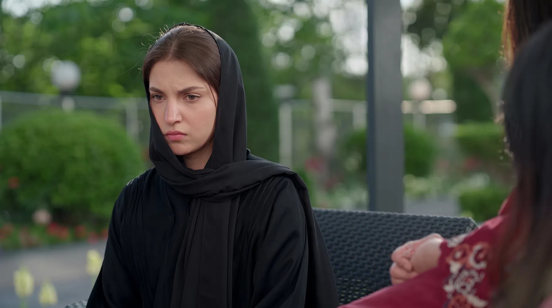 An intense close-up shot captures Pakistani actress Dananeer Mobeen portraying a moment of deep solemnity and emotional distress, her focused expression conveying heavy grief and determination. Clad in a traditional black abaya with a matching hijab pulled snugly over her head, she sits outdoors against a beautifully blurred background of lush green foliage and gardens. To the side, the soft-focus profile of another individual in a patterned maroon outfit hints at a tense, interpersonal conversation unfolding in an elegant open-air setting.