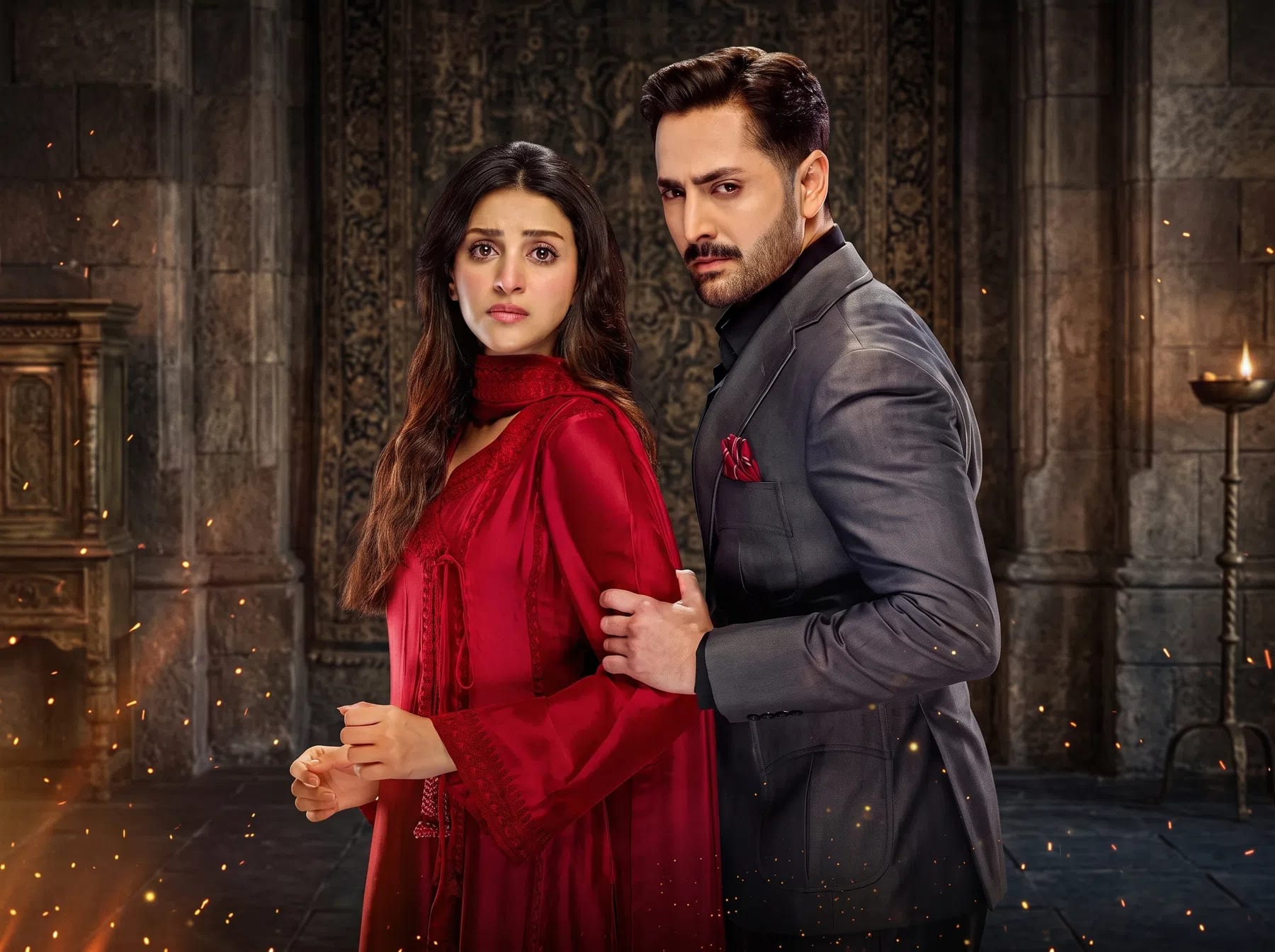 An intense promotional poster features Pakistani actors Danish Taimoor and Anmol Baloch standing close together against a dark, dramatic stone backdrop. On the left, Baloch is captured from the waist up, dressed in a vibrant red traditional outfit with her long dark hair falling over her shoulders, her expression one of worry or concern. On the right, Taimoor stands in profile, looking directly at the camera with a stern, focused gaze. He is wearing a sharp, dark grey tailored suit jacket over a black shirt, with a red pocket square matching her attire. His hand is placed firmly on her arm, suggesting a protective or controlling stance. The atmosphere is elevated by warm lighting, flying sparks, and a classical stone archway with an ornate screen behind them. Drama seriel Nashtar, image by PakistaniArt.