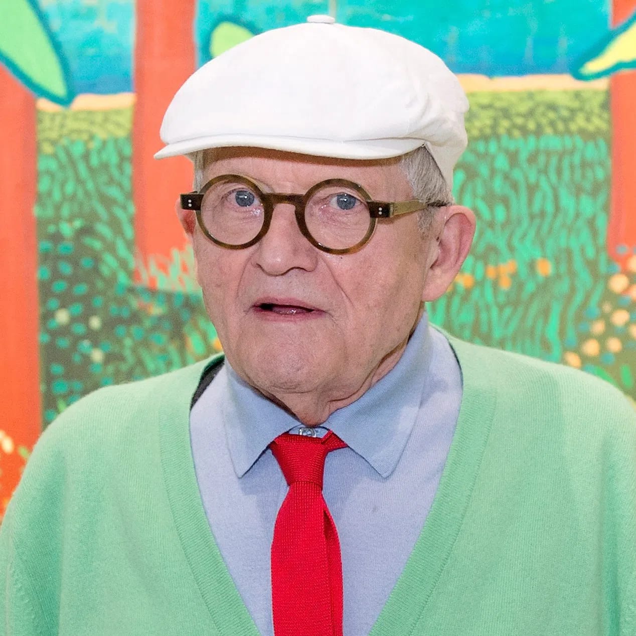 A close-up photograph of British artist David Hockney wearing a white flat cap, round horn-rimmed glasses, a pastel green cardigan, a light blue dress shirt, and a vibrant red knit tie. He stands in front of one of his colorful landscape paintings depicting red trees and stylized green leaves.
