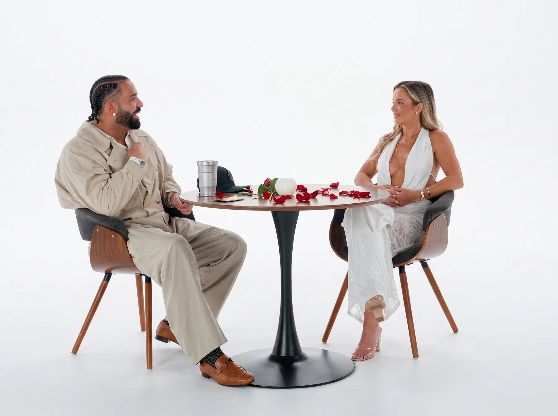 The image captures a professionally lit studio setting featuring Canadian rapper and global superstar Drake sitting across a round, dark-wood bistro table from a woman during a speed dating sequence. The table is distinctively decorated with scattered red rose petals. Drake is positioned on the left side of the frame, styled in matching loose-fitting, beige or tan utility streetwear pants and a matching jacket over a white undershirt. His hair is neatly braided into cornrows, and he is wearing brown leather loafers with white socks, leaning slightly forward with a broad smile while gesturing with his right hand toward his companion.Seated opposite him on the right is a woman with blonde hair, wearing a deeply plunging, halter-neck white dress featuring a sheer lace overlay skirt. Both individuals are sitting in modern mid-century styled armchair seats equipped with dark grey padded cushions and curved walnut wood frames supported by tapered dowel legs. The backdrop is entirely flat and seamless white studio space, creating a stark, high-contrast visual environment that draws all attention to the interactive date setup at the center.