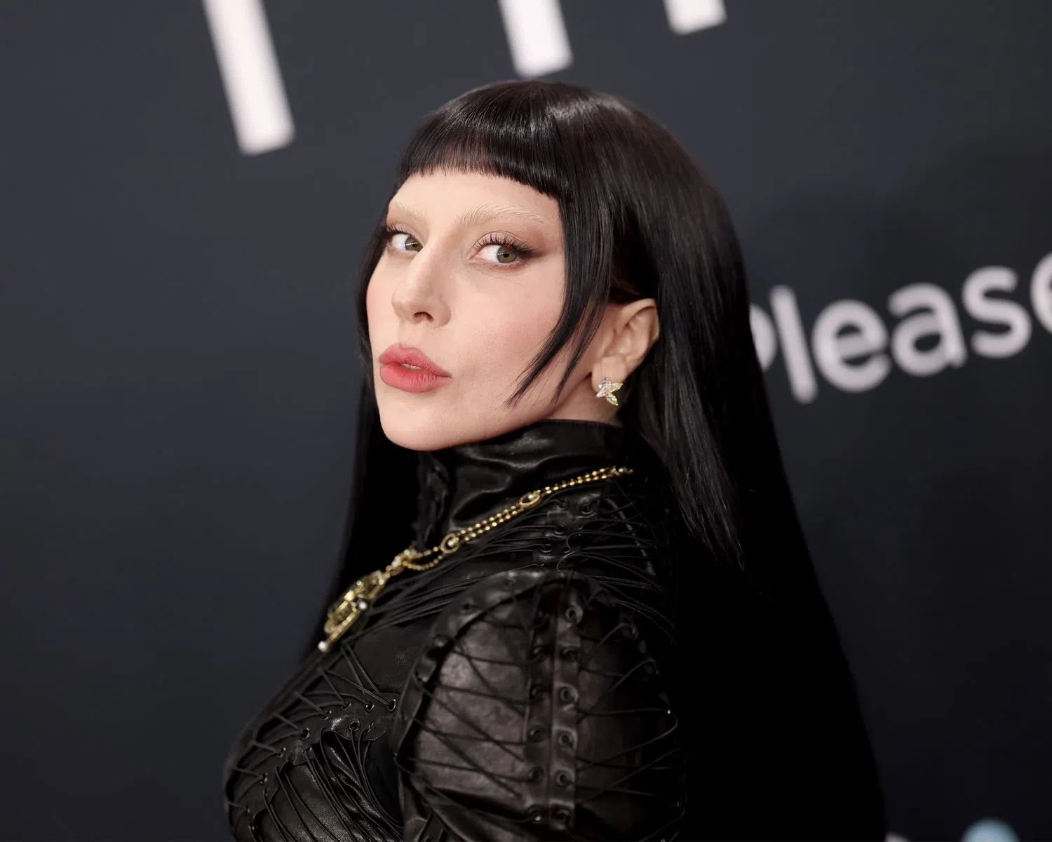 Lady Gaga delivers a striking visual statement on the red carpet with a high-drama beauty look defined by ultra-short micro bangs, long raven-black hair, and completely bleached eyebrows. Her complexion is flawless and luminous, paired with a sharp, subtle winged eyeliner that draws focus to her eyes, and a soft, matte rose lip color. She wears a high-necked, avant-garde black textured garment featuring intricate lacing details, accessorized with a statement gold chain necklace and delicate, light-colored earrings that catch the light, all set against a dark backdrop with blurred event signage.