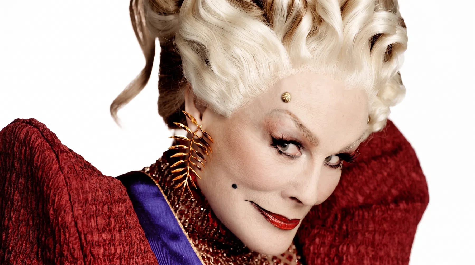 Glenn Close commands the frame with a wicked, theatrical intensity as Drusilla Sickle, showcasing a striking villainous look complete with an elaborate platinum blonde wig styled in structured vintage waves, dramatic arched eyebrows accented by a gold micro-stud, and a sharp, glossy red lip featuring a defined dark outline. Her piercing gaze is directed sharply to the side while wearing an oversized, structural red textured collar that frames her face, paired with a matching gold fern-like statement earring that perfectly complements her sinister, aristocratic aesthetic. Glenn Close transformed as the character Drusilla Sickle in The Hunger Games: Sunrise on the Reaping.