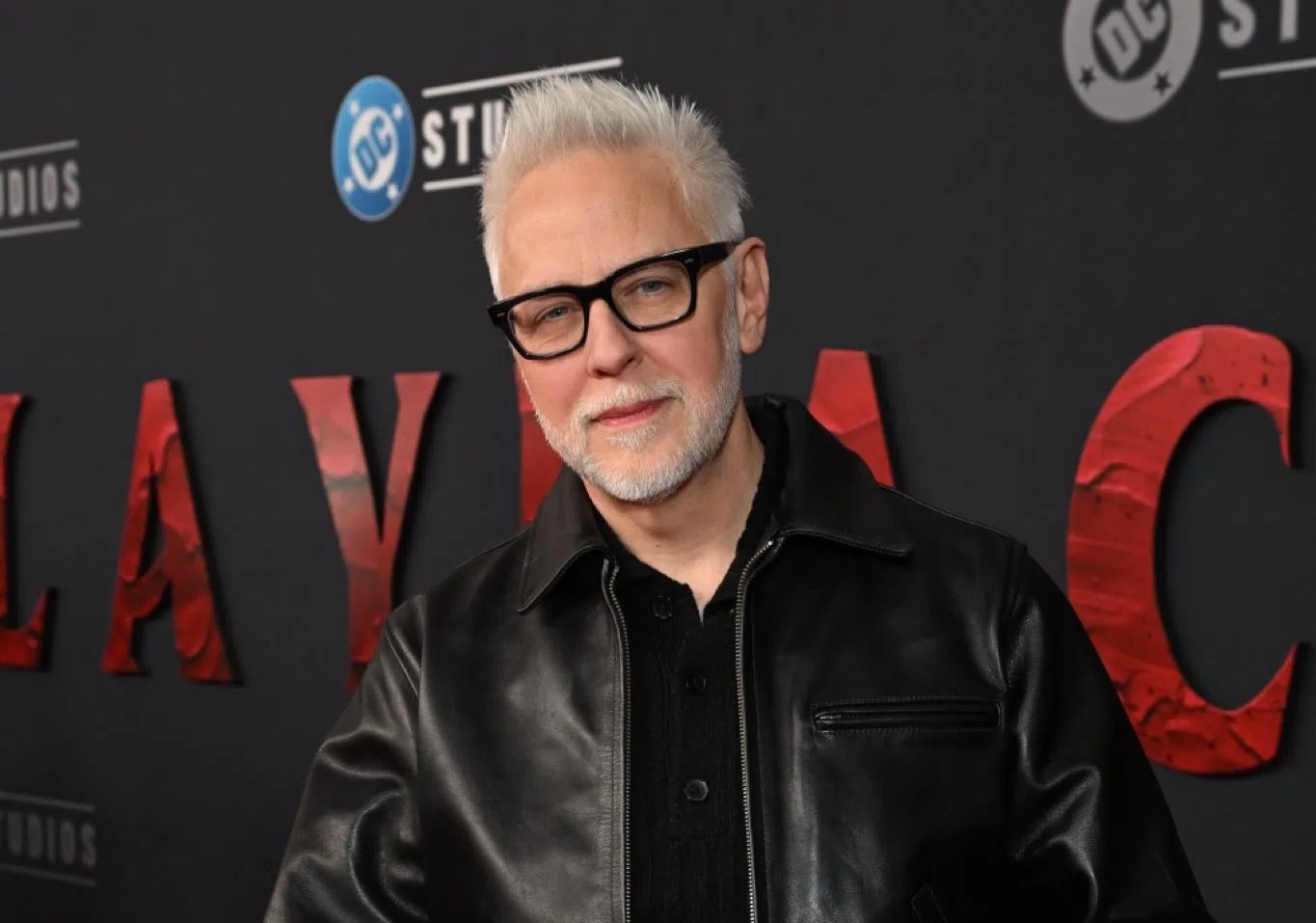 The image shows filmmaker James Gunn attending the premiere of "Clayface." Gunn is captured from the chest up, positioned slightly off-center to the right, against a dark gray backdrop featuring the text "CLAYFACE" in large, textured red capital letters, alongside the DC Studios logo. He is smiling faintly, looking directly at the camera, and has short, styled white hair with a light gray beard and mustache. He is wearing thick-rimmed black eyeglasses, a black collared button-down shirt, and a classic black leather jacket with a pointed collar. The lighting highlights the texture of his jacket and hair, creating a sharp contrast against the dark background.