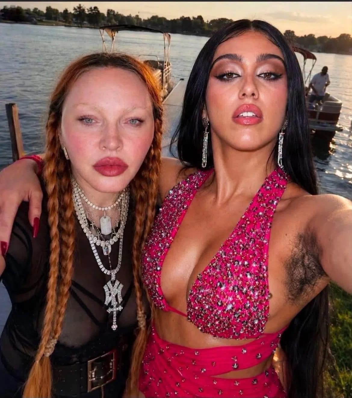 The image shows Madonna and her daughter, Lourdes Leon, standing closely together on a dock beside a body of water at sunset. Madonna, on the left, features long, reddish-blonde braided hair, bleached eyebrows, prominent pink blush, and bold red lipstick. She is wearing a black mesh top, a wide buckled belt, and layered silver chain necklaces, including a large cross pendant. Lourdes Leon, on the right, has long, straight black hair, dramatic eye makeup, and glossy lips. She wears a vibrant, embellished pink halter top with matching wrap detailing, raising her arm to take the selfie and naturally revealing her unshaved underarm. In the background, calm water stretches out with a pontoon boat docked nearby and trees lining the distant shore under a soft evening sky.