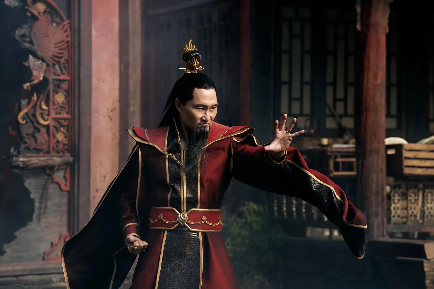 In this promotional still from the live-action adaptation of Avatar: The Last Airbender, actor Daniel Dae Kim perfectly embodies the tyrannical and ruthlessly driven ruler of the Fire Nation, Fire Lord Ozai. Clad in his highly detailed, regal crimson-and-black imperial robes accented with ornate gold trim, Ozai strikes an aggressive, commanding firebending stance with an outstretched open palm. His long black hair is partially pulled back into a traditional topknot secured by a sharp, flame-styled gold hairpiece, complementing his distinctive long, sharp goatee. The intense, calculating glare across his face highlights his absolute desire for total world conquest, captured in a dimly lit, traditional East Asian architectural courtyard that echoes the oppressive, industrial aesthetic of the Fire Nation Palace.