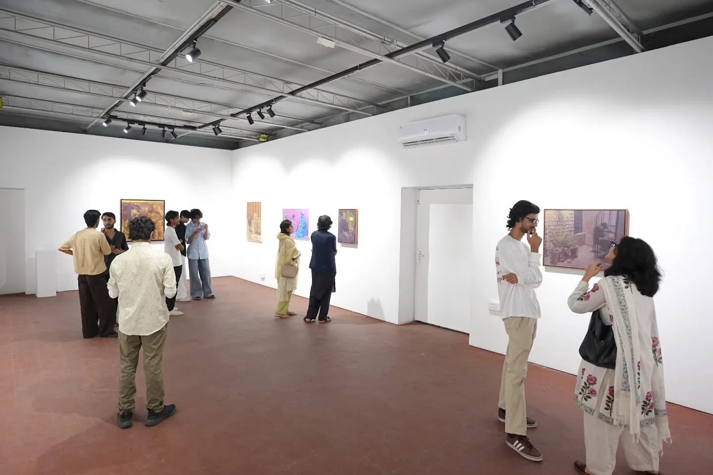 An indoor art gallery setting shows an exhibition titled 'Of Seeing Men: Everyday Masculinities' at ZQ Gallery, where multiple visitors are spread across a spacious room with white walls and a reddish-brown floor. Art enthusiasts, dressed in casual and traditional attire, stand in small groups or individually, closely observing various framed paintings hung at eye level along the walls. The gallery features a minimalist industrial ceiling with exposed metal trusses, track lighting pointing toward the artworks, and an air conditioning unit mounted high on the wall, creating a bright and contemplative atmosphere for the attendees.