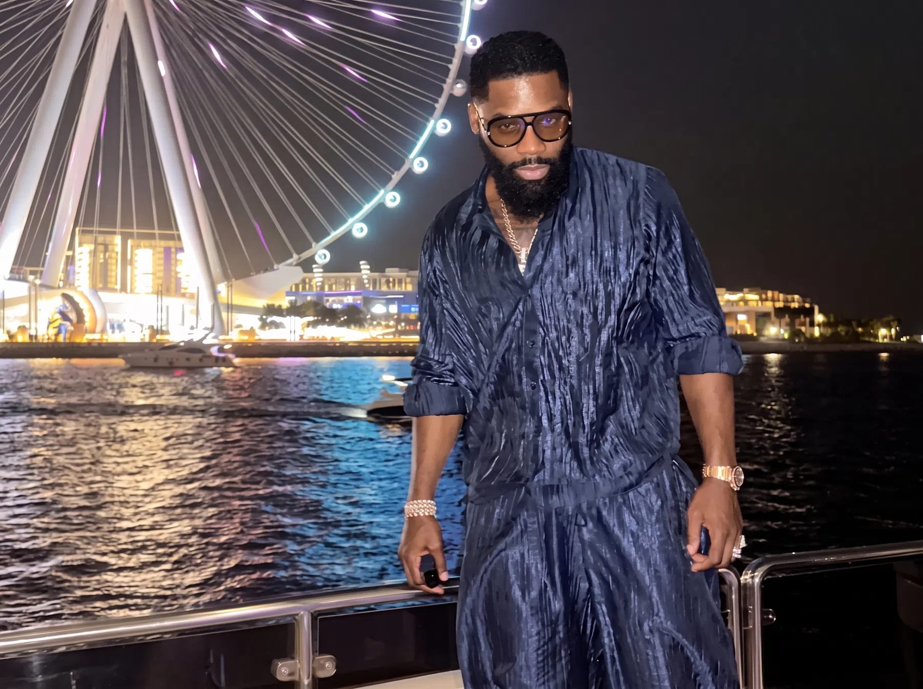 The image features Igho "Tiny" Ubiribo posing confidently on a boat at night, set against the vibrant backdrop of a massive, illuminated Ferris wheel—the Ain Dubai—stretching into the dark sky. He is dressed in a stylish, matching dark navy blue plissé or crinkled two-piece shirt and pants set that subtly catches the surrounding city lights. Accessorizing with large, tinted sunglasses, a thick beard, a classic luxury watch, and a bracelet, he leans casually against the boat's metallic railing. The dark, shimmering water in the foreground reflects the brilliant white and gold neon glows from the observation wheel and the distant coastal skyline, creating a sophisticated and upscale nocturnal atmosphere.