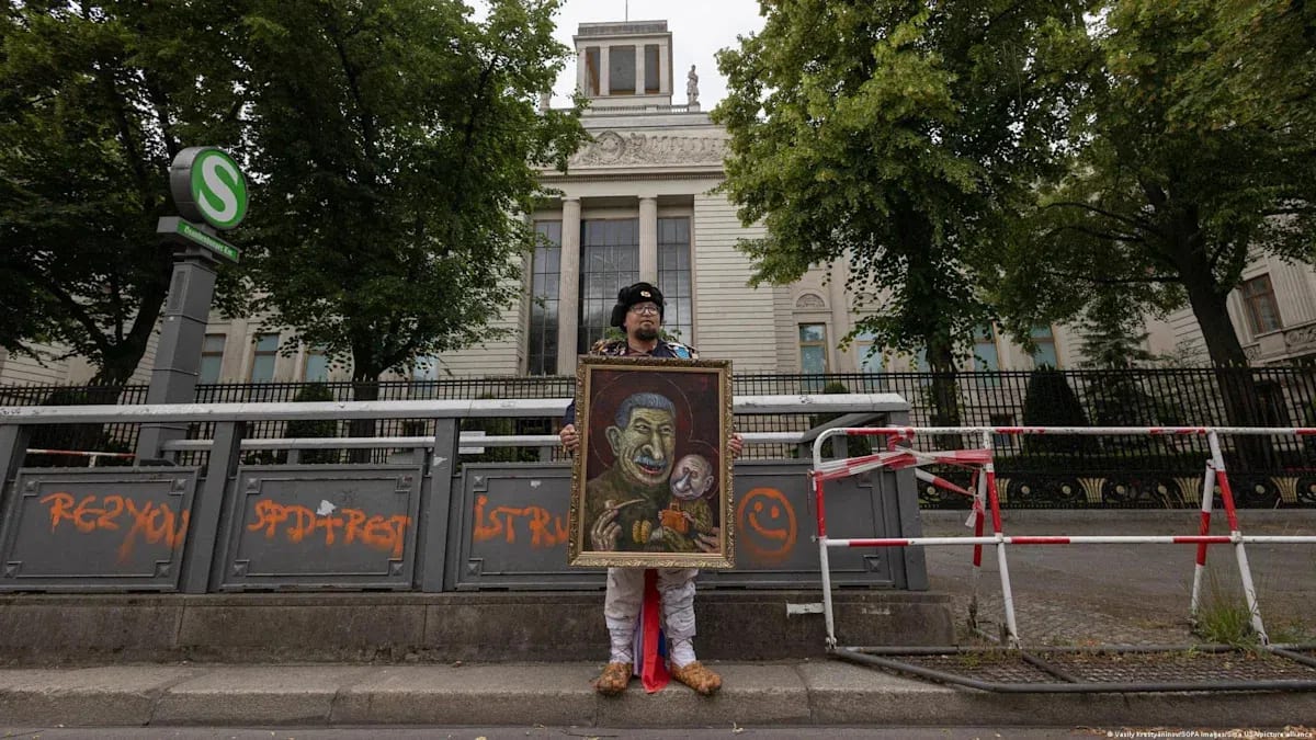 Russian artist and activist Semyon Skrepetsky stands on a city sidewalk in front of a grand, multi-story building, holding a large framed painting that depicts a caricature of two figures, one smoking a pipe. The individual wears a distinctive dark ushanka hat, glasses, patterned clothing, and light-colored pants draped with a Russian flag. The scene unfolds behind a temporary metal barricade sprayed with orange graffiti, next to an entrance marked by a green "S" transit sign.