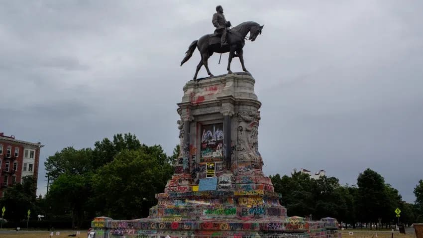 The graffiti-covered statue of Confederate General Robert E. Lee catches the early morning light on June 15, 2020 at Monument Avenue in Richmond, Virginia. An equestrian bronze statue of Confederate General Robert E. Lee sits atop a massive, ornate stone pedestal covered entirely in colorful graffiti, tags, and protest art, captured under an overcast, cloudy gray sky. The monument dominates the center of the frame, surrounded by green trees in the background and a brick building visible on the far left.