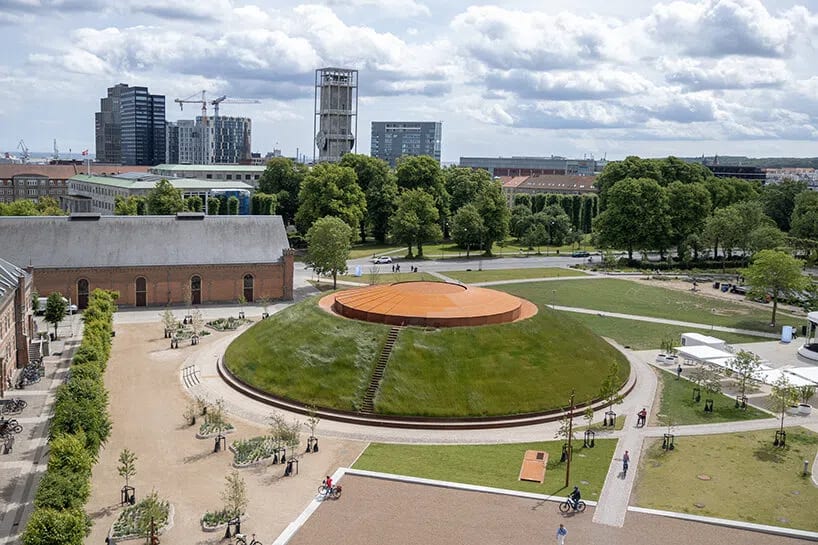The outdoor view of James Turrell's monumental 40-metre-wide Skyspace, titled "As Seen Below – The Dome", presents itself as a low, grass-covered circular mound rising from the public park directly beside the ARoS Aarhus Art Museum in Aarhus, Denmark.Designed in collaboration with Schmidt Hammer Lassen Architects, the exterior of the installation is integrated into the surrounding landscape as a minimalist architectural intervention.Exterior Architecture and Visual DynamicsThe Grassy Mound: From the outside, the massive subterranean concrete dome is hidden beneath a convex, turf-covered hill that blends directly into the museum campus parkland.The Dark Oculus: At the very apex of this green mound sits a sharp, 6-metre-wide open circular aperture (oculus). From an aerial or elevated perspective, this opening appears as a deep, geometric cut that frames the sky.Dialogue with "Your rainbow panorama": The low, subterranean nature of the Skyspace acts as an intentional structural counterpoint to Olafur Eliasson’s famous Your rainbow panorama, the vibrant, elevated glass walkway that sits high on the roof of the main ARoS building. While Eliasson's work projects visitors outward to view the city through color, Turrell's dome pulls the landscape inward, framing the Danish sky as a localized, living artwork.