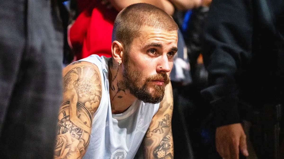 A close-up shot captures Justin Bieber from the chest up, leaning slightly forward with a pensive and focused expression while looking toward the right. He features a buzz-cut hairstyle, a well-groomed full beard, a mustache, a small hoop earring in his right ear, and a subtle nose stud. He is wearing a light blue sleeveless jersey, which fully exposes his arms and neck to reveal extensive, intricate black-and-grey ink tattoos, including an angel and a clock face design on his right shoulder and bicep. The foreground and background are softly blurred, showing glimpses of dark clothing from individuals standing nearby, creating a shallow depth of field that keeps the central focus entirely on his contemplative demeanor.
