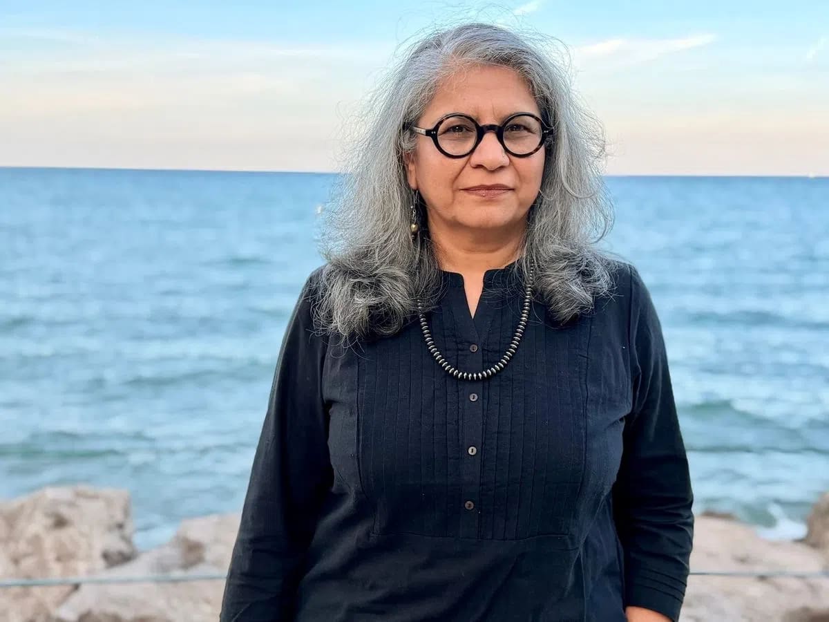 A medium close-up shot captures Dr. Savneet Talwar standing outdoors against a backdrop of a calm, blue sea meeting a light sky at the horizon line. She has shoulder-length, wavy grey hair parted to the side, and she wears round, thick-rimmed black glasses. Her expression is composed and neutral as she looks directly toward the camera. She is dressed in a dark black, long-sleeved button-up shirt featuring subtle vertical pleated detailing on the chest, complemented by a simple dark metallic beaded necklace. In the lower-left foreground, blurred rocky terrain lines the shore, establishing an outdoor coastal setting.