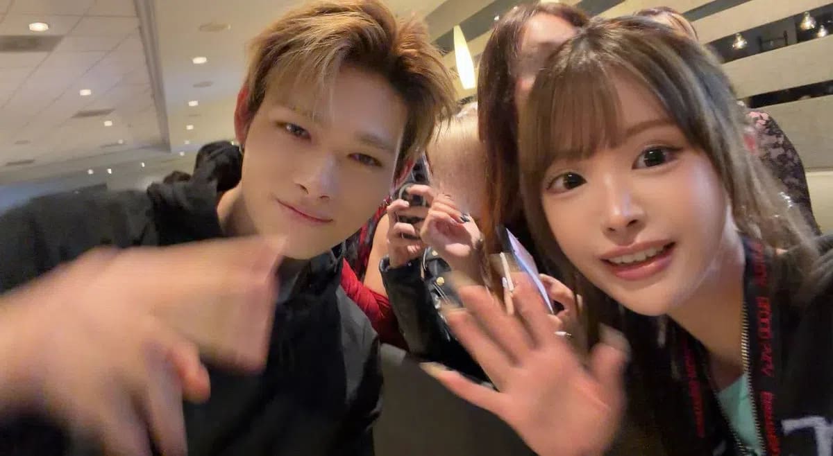 A slightly blurry, close-up selfie captures Ni-Ki from the K-pop group ENHYPEN alongside a female fan as they both wave toward the camera. On the left, Ni-Ki leans into the frame with styled brownish-blonde hair, wearing a casual black hooded jacket while extending a motion-blurred hand forward in a greeting gesture. To the right, the fan smiles warmly, sporting straight brown hair with bangs and holding up her hand to wave, with her lanyard visibly draped around her neck. In the background, out-of-focus figures of other people and the warm, bright overhead lighting of an indoor venue or eatery are visible, creating a candid and energetic atmosphere.
