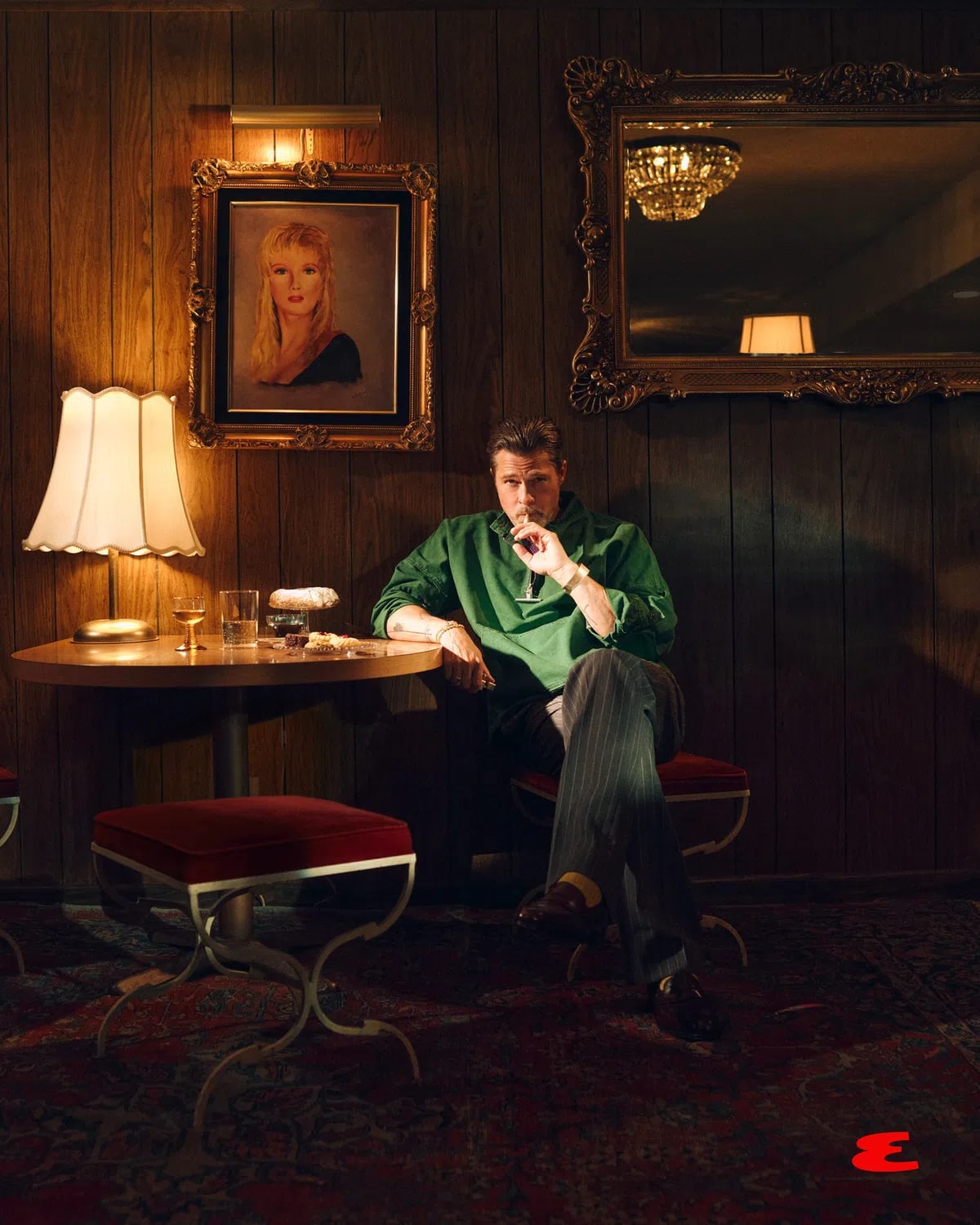A visually rich, cinematic photograph captures actor Brad Pitt seated in a retro, wood-paneled room with a moody, warm ambiance. He wears a vibrant green pullover, pinstriped trousers, and brown loafers while holding a cigar near his mouth. He sits cross-legged next to a small wooden table illuminated by a scalloped-edge lamp, displaying glasses and pastries. Behind him, an ornate gold-framed portrait of a blonde woman hangs on the dark wood wall, and a large matching mirror reflects a glowing chandelier. The setting features a classic Persian rug on the floor and a plush red stool in the foreground, evoking a nostalgic, mid-century lounge aesthetic.