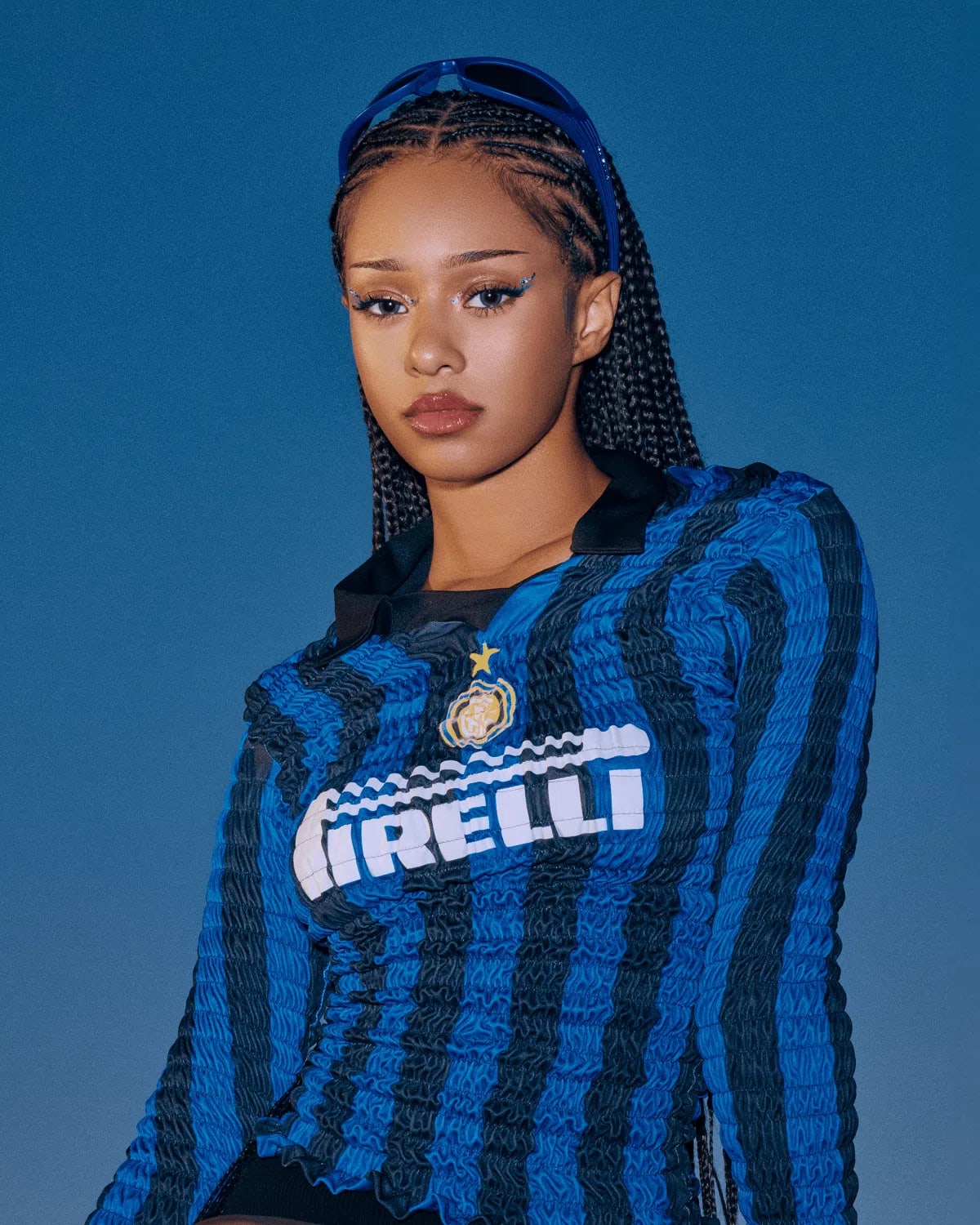 The image shows a portrait of model and content creator Manon Bannerman set against a solid blue background, styled in a sporty, Y2K-inspired aesthetic. She wears an Inter Milan football shirt featuring vertical blue and black stripes, a black collar, and the white "Pirelli" sponsor logo across the chest, uniquely updated with a crinkled, textured popcorn fabric. Her hair is styled in neat, symmetrical cornrow braids extending to the back, with blue-framed sunglasses resting on top of her head. Her makeup is sharp, highlighting soft brown lipstick, winged graphic eyeliner adorned with small silver metallic accents near the eyes, and well-defined brows. A portrait showcases Manon Bannerman wearing a vintage-style Inter Milan soccer jersey modified into a smocked, texture-rich long-sleeve top featuring the classic white and yellow Pirelli front sponsor logo against vertical blue and black stripes. She is styled with neat, long box braids drawn straight back, dark sunglasses propped up on her head as a hairband, subtle cat-eye eyeliner, and neutral lip gloss, all set against a solid deep blue background.