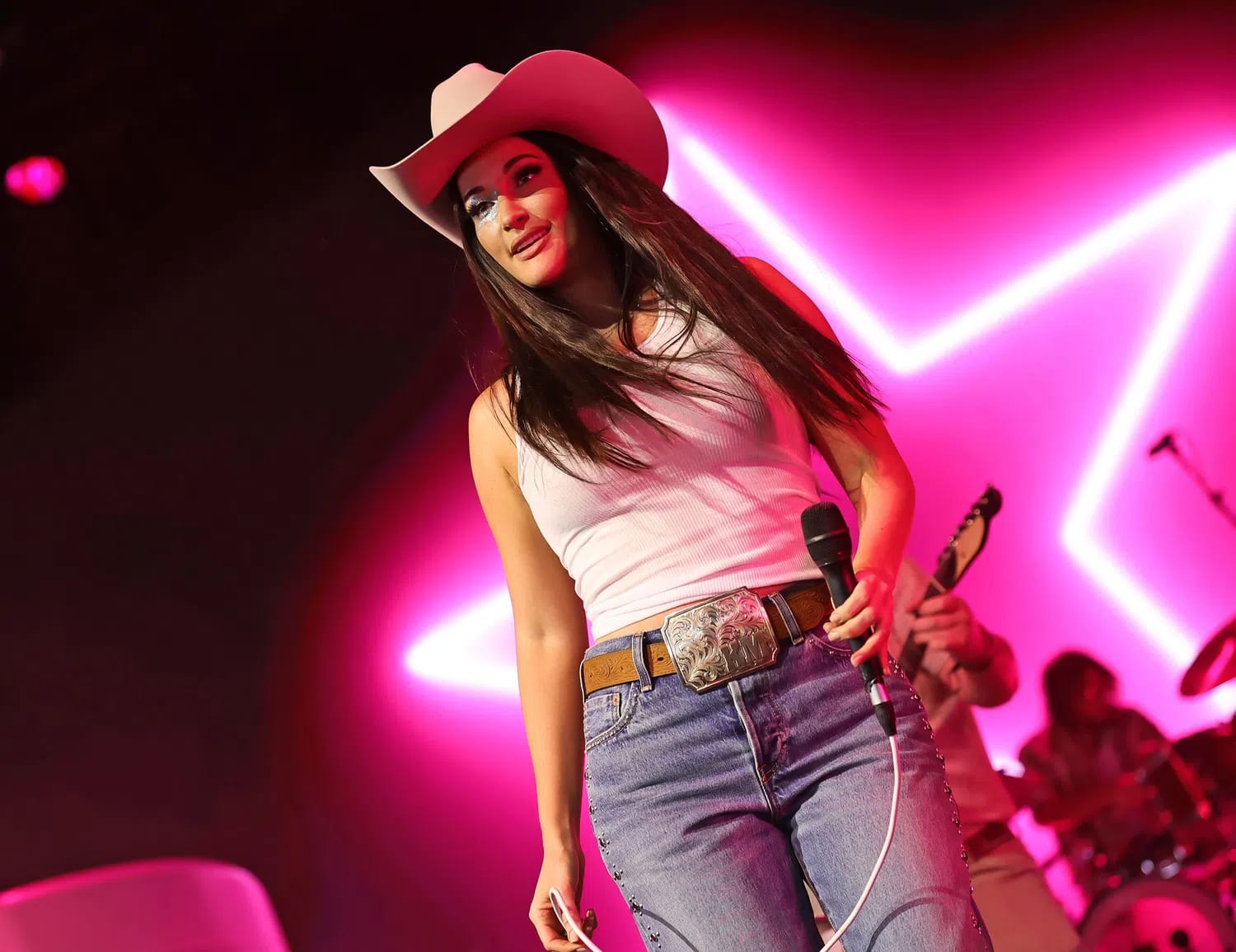 A medium shot captures singer Kacey Musgraves performing on stage under vibrant pink neon lighting. She is wearing a classic white ribbed tank top, medium-wash blue jeans with metallic studded detailing down the sides, and a brown leather belt featuring a large, ornate silver Western buckle. Her look is completed with a light-colored cowboy hat and bold, shimmering blue eyeshadow. She holds a black microphone in her left hand, moving forward on stage with a smiling expression, while a large neon star glows brightly in the background behind her and her band members.