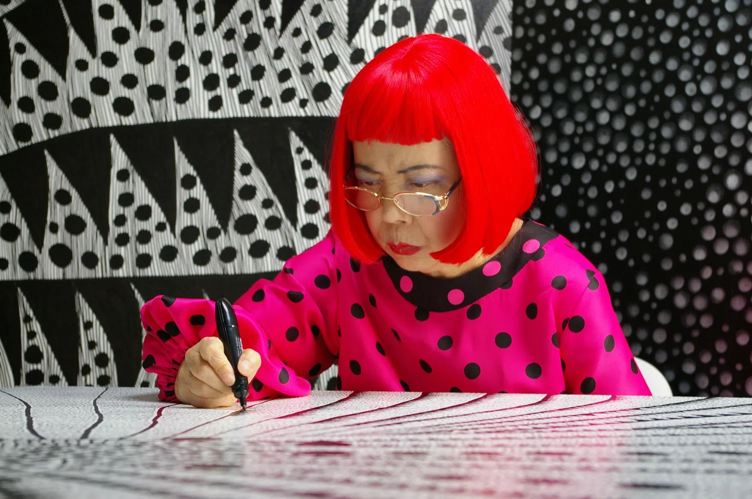 The image shows the famous contemporary Japanese artist Yayoi Kusama working diligently at a desk, deeply focused on drawing intricate patterns with a black marker on a large canvas. She is instantly recognizable by her vibrant, bright red bob haircut with straight-across bangs, gold-rimmed reading glasses, and dark eyeshadow. She wears a striking hot pink blouse covered in large black polka dots, featuring a black collar and long sleeves with gathered, elasticated cuffs. Her hand holds the marker firmly as she guides it across a surface densely covered in her signature fine lines and repetitive dot motifs. The background behind her is a visually complex, high-contrast environment filled with her artwork, displaying large black and white geometric shapes, alternating fields of dense polka dots, and vertical patterns that create an immersive, hallucinatory atmosphere characteristic of her iconic style.A high-contrast photograph of artist Yayoi Kusama in her signature bright red bob and polka-dot blouse creating detailed artwork.