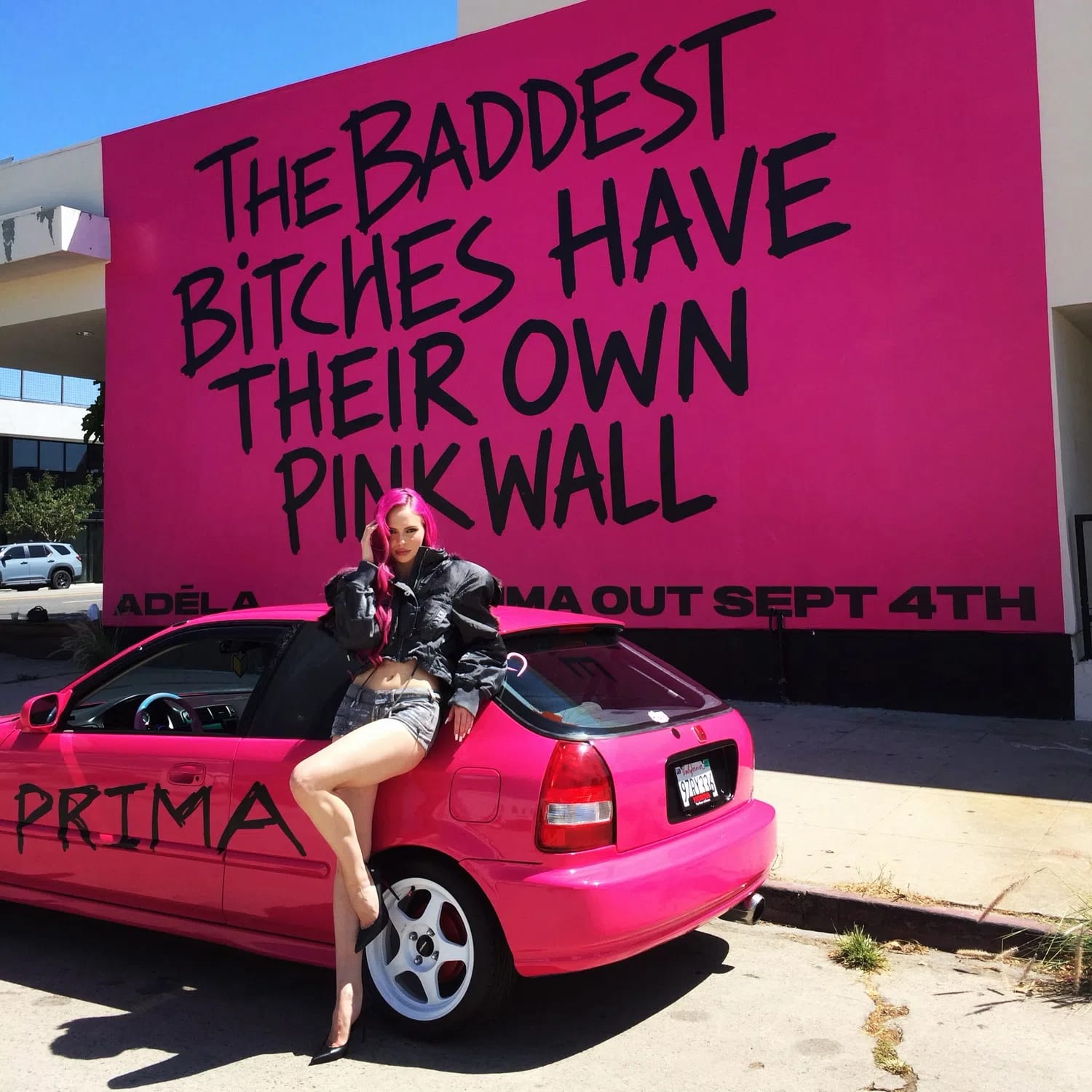 The image shows a vibrant promotional scene featuring artist ADÉLA posing on a custom hot pink car in front of the iconic Paul Smith Pink Wall on Melrose Avenue in Los Angeles. The wall features a bold, graffiti-style black inscription that reads, "THE BADDEST BITCHES HAVE THEIR OWN PINK WALL," alongside text underneath announcing her project "PRIMA OUT SEPT 4TH." ADÉLA matches the aesthetic with hot pink streaks in her hair, denim shorts, and a black puffer jacket, while sitting on a pink hatchback that has "PRIMA" spray-painted across the side door.
