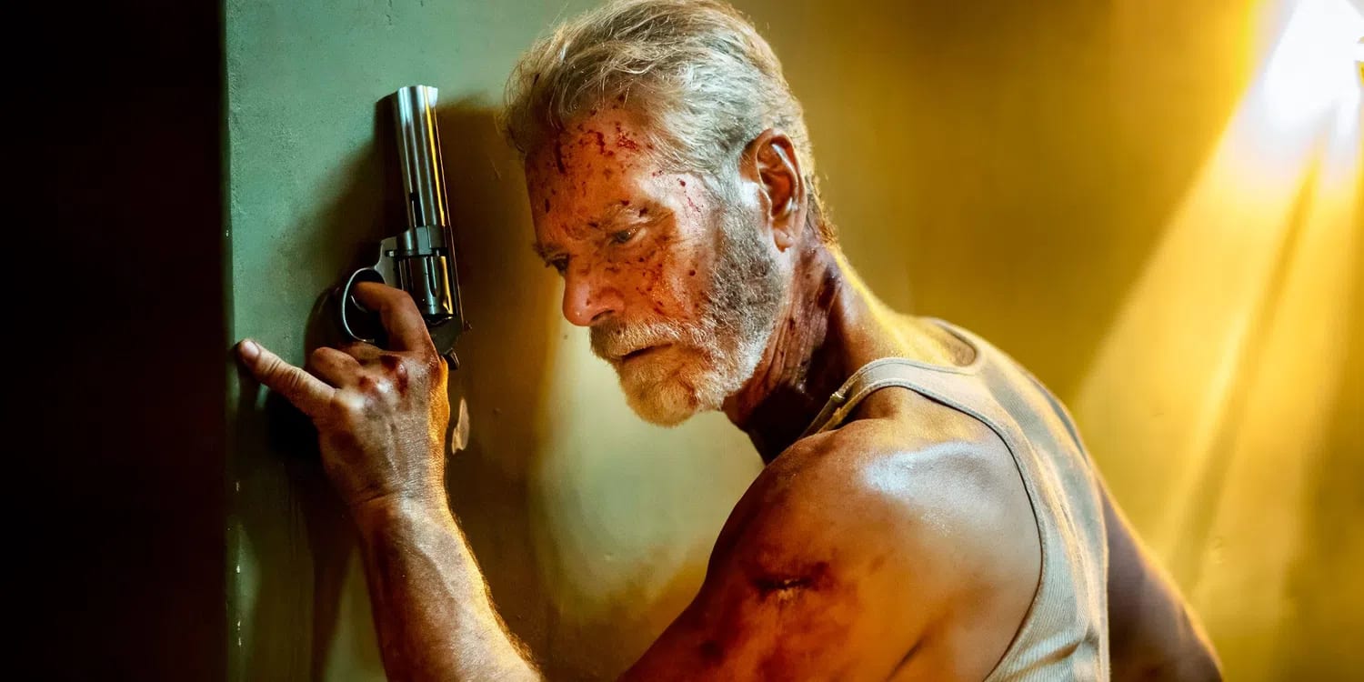 Stephen Lang stars as the formidable Norman Nordstrom, a blind military veteran navigating intense combat in a dimly lit, sun-streaked interior hallway. In this high-contrast cinematic shot from the horror-thriller film Don't Breathe 2, Nordstrom leans heavily against a dark wall, his expression a mix of fierce concentration and physical exhaustion. His face, arms, and white tank top are heavily stained with blood and grime, highlighting the brutality of the conflict. He holds a large, silver revolver upward with a steady grip, his finger resting near the trigger as he listens intently for approaching threats. A warm, harsh light streams from the right background, cutting through the shadows and illuminating the dust in the air, emphasizing the gritty and suspenseful atmosphere of his isolated environment.