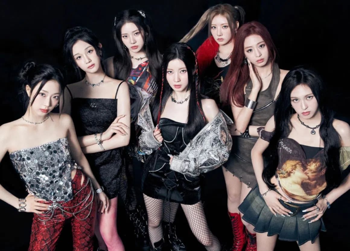 An group of seven young women pose together closely against a dark background, showcasing an edgy, modern fashion aesthetic. On the far left, a woman with dark hair tied back wears a strapless, glittering silver-and-black top paired with textured red trousers and a silver chain belt. Next to her, a woman in a black spaghetti-strap dress stands with her arms crossed, sporting a matching dark necklace. Behind them, a woman with long dark hair and a plaid-accented top looks forward. In the center, a woman with braided sections in her dark hair wears a black strapless dress under an oversized metallic silver jacket, paired with fishnet stockings. To her right, a woman with high pigtails and a dark top with a red fuzzy collar glances slightly sideways. Next to her, a woman with long red-toned hair poses with her hand to her cheek, dressed in an olive-green sleeveless top and matching skirt. On the far right, a woman with long, wavy dark hair wears a top featuring a classic art print of cherubs, paired with a pleated grey denim miniskirt. All members present a confident, synchronized expression toward the camera under cool-toned studio lighting.