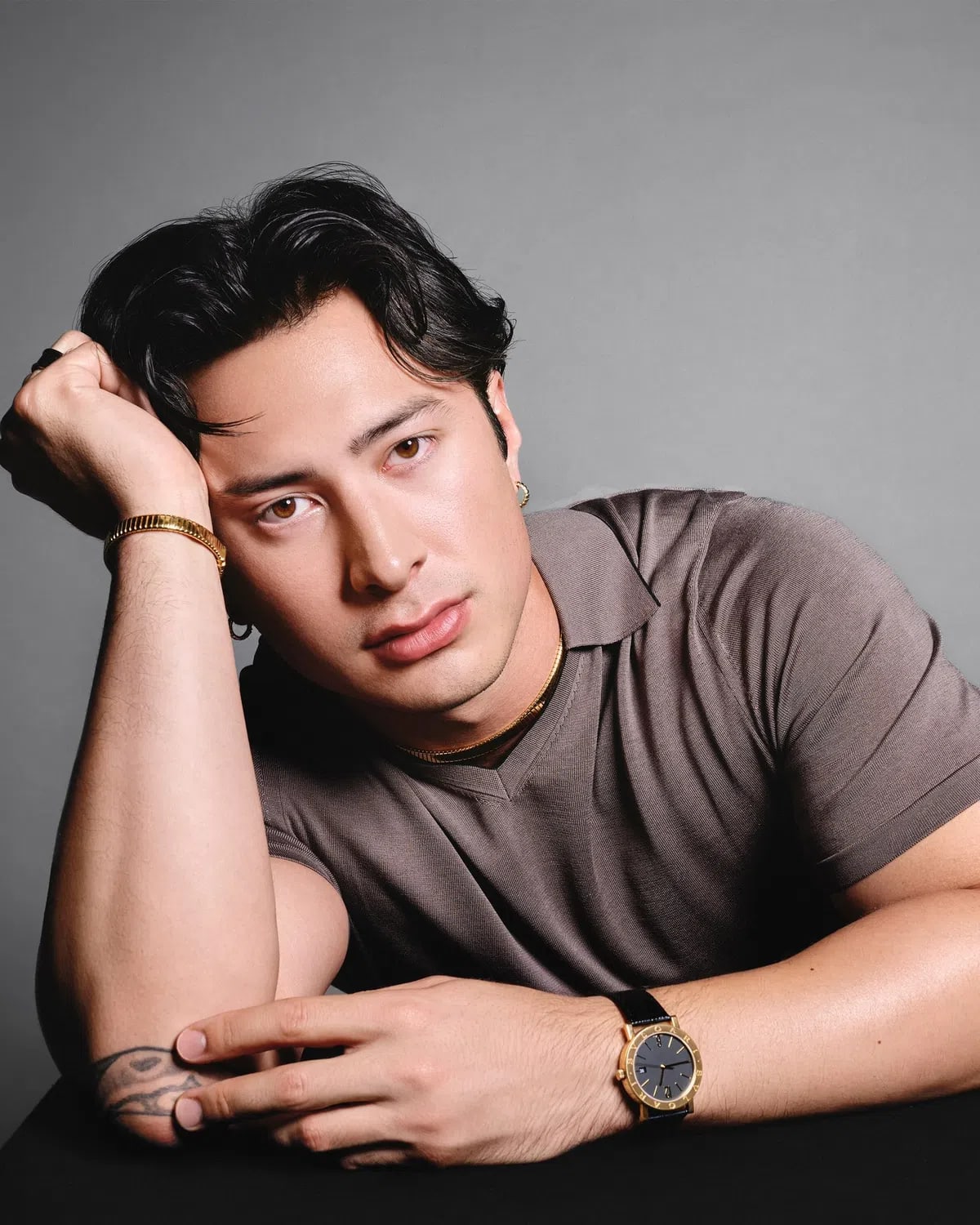 The image captures a striking studio portrait of Hudson Williams, looking directly into the camera with a calm, introspective expression while leaning forward against a dark surface. His dark, wavy hair is parted down the middle and styled elegantly, framing a sharp face highlighted by warm lighting against a neutral, slate-gray backdrop. He is wearing a short-sleeved, taupe-brown knit polo shirt with a fine textured ribbed collar, showing a partial tattoo on his right inner forearm. His right hand rests against his temple, showcasing an ornate gold bracelet and small gold hoop earrings, while his left arm rests horizontally across the bottom of the frame, drawing attention to a luxury Bvlgari watch with a gold case and a black leather strap.