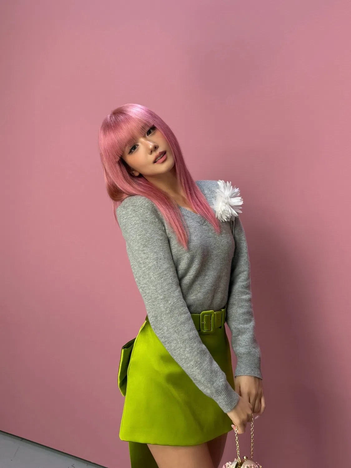 A young woman with vibrant pink hair and bangs poses against a pink background, wearing a grey long-sleeved sweater with a large white flower pin on the shoulder and a structured lime-green belted miniskirt while holding a small gold chain handbag. The image showcases a vibrant and stylish fashion portrait set against a solid, matte pink background. A young woman with long, straight pink hair and a clean fringe leans gracefully to her left, looking directly into the camera. She wears a long-sleeved, grey knit sweater adorned with a large, white fabric flower pinned to her left shoulder. The sweater is tucked into a bright, structured lime-green miniskirt featuring a matching fabric belt with a square buckle. The skirt includes a structured panel or bow detailing extending down the side and back. Her left hand rests near the belt, while her right hand gently holds a small, embellished gold chain purse.