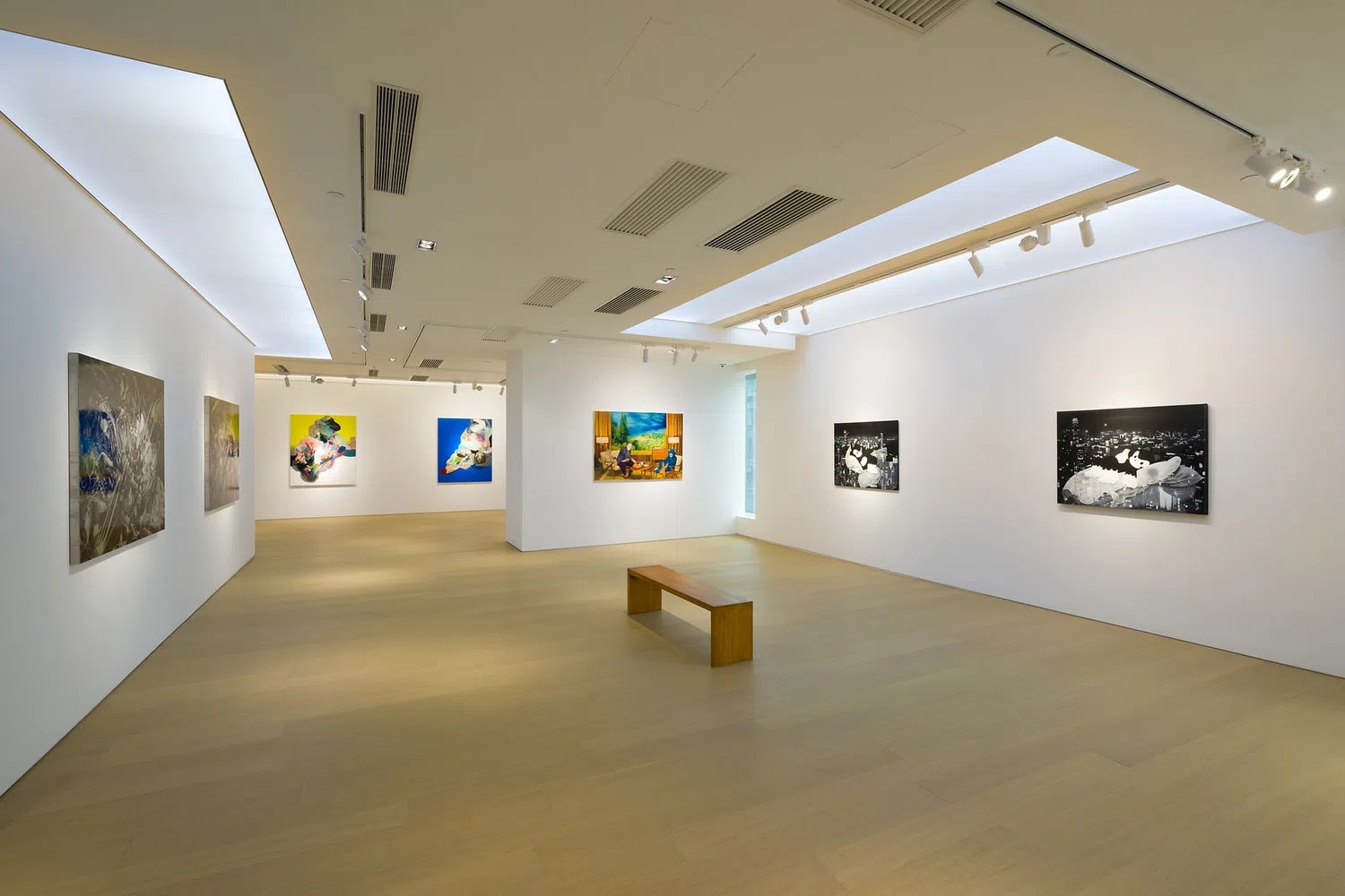 Tang Contemporary Art Gallery exhibition space featuring contemporary paintings on display under bright gallery lighting. An expansive, minimalist art gallery interior features light wood floors and clean white walls illuminated by modern recessed ceiling lights and track lighting systems. The wide-angle perspective showcases a spacious exhibition hall where vibrant contemporary paintings are spaced out across the walls, leading the viewer's eye toward a central wooden bench and an adjacent hallway that extends deeper into the gallery.
