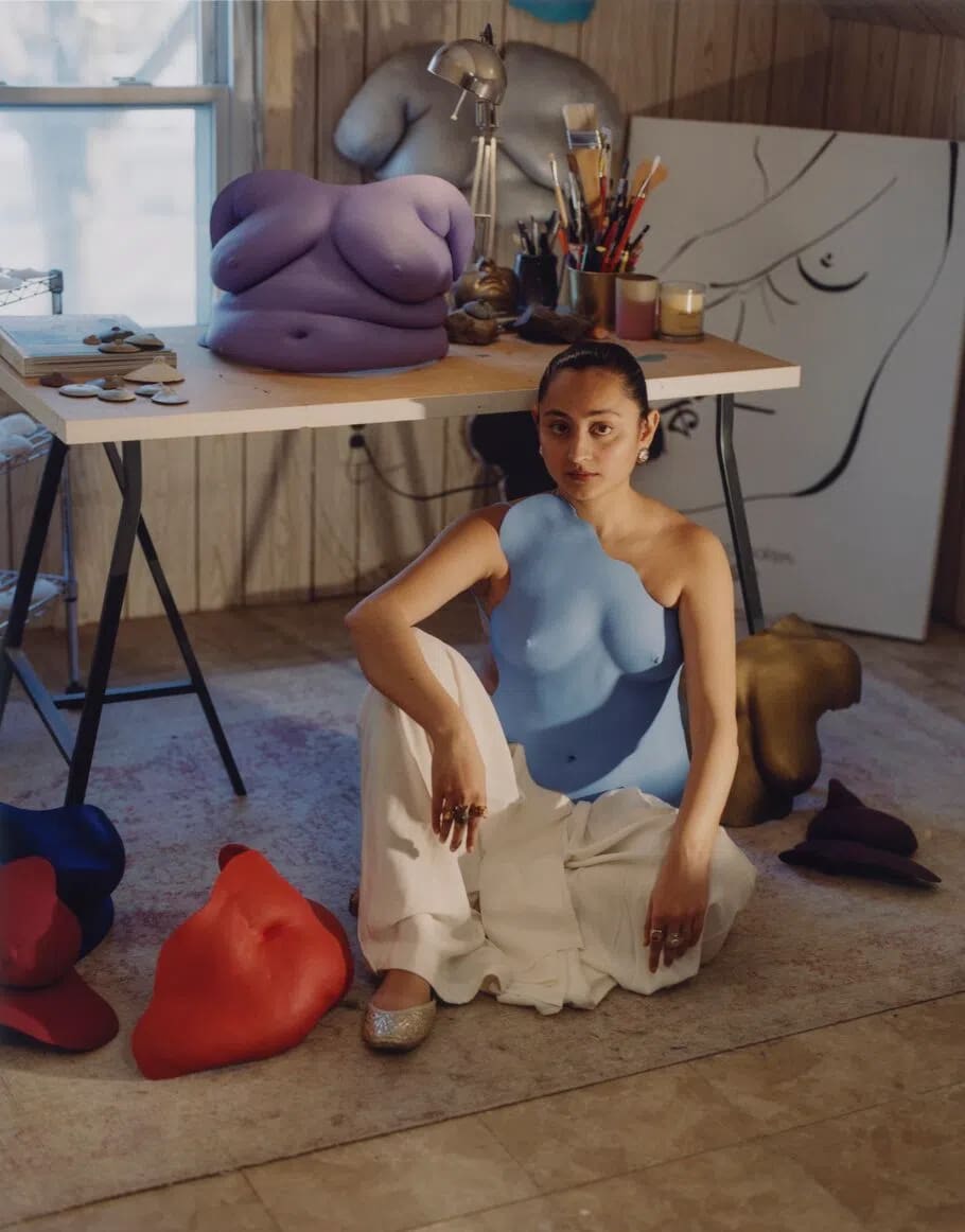 In this portrait, the Pakistani artist and fashion designer Misha Japanwala sits on the floor of her art studio surrounded by several of her signature, handcrafted body-cast sculptures. She is positioned centrally, looking directly at the camera while wearing white, wide-leg trousers and a striking, asymmetrical sky-blue resin breastplate molded to her torso. Behind her stands a light-wood work desk covered with art supplies, including a jar of paintbrushes and candles, and topped by a large, lavender-colored sculptural torso. Propped against the wall behind the desk is a large canvas featuring a minimalist black-and-white calligraphic silhouette drawing. Scattered around her on the floor are additional sculpted anatomical forms in bold shades of red, dark blue, gold, and dark purple, highlighting her unique creative practice that explores themes of bodily autonomy and the deconstruction of shame.
