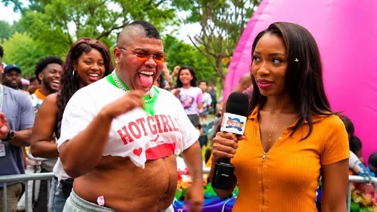 The image showcases a viral comedy sketch from digital creator and comedian Druski, titled ["Kid Stars grow up & be so Unpredictable," featuring his parody character Zion. In this scene, Druski plays a eccentric, unpredictable adult version of a fictional former Disney Channel icon, parodying the wild paths of child actors navigating fame after their childhood shows end. Dressed in an altered, midriff-baring white t-shirt reading "HOT GIRL" alongside distinctive dyed orange glasses and a mohawk hair strip, he executes chaotic, high-energy antics while sticking his tongue out. Beside him stands an actress portraying a field reporter holding a news microphone, dressed in a ribbed mustard-orange zip-up top. The background features a large pink inflatable tent and festival crowds at an outdoor event, capturing the humorous, satirical tone of the piece that many online viewers immediately linked to interviews and public figures like Orlando Brown and Raven-Symoné.