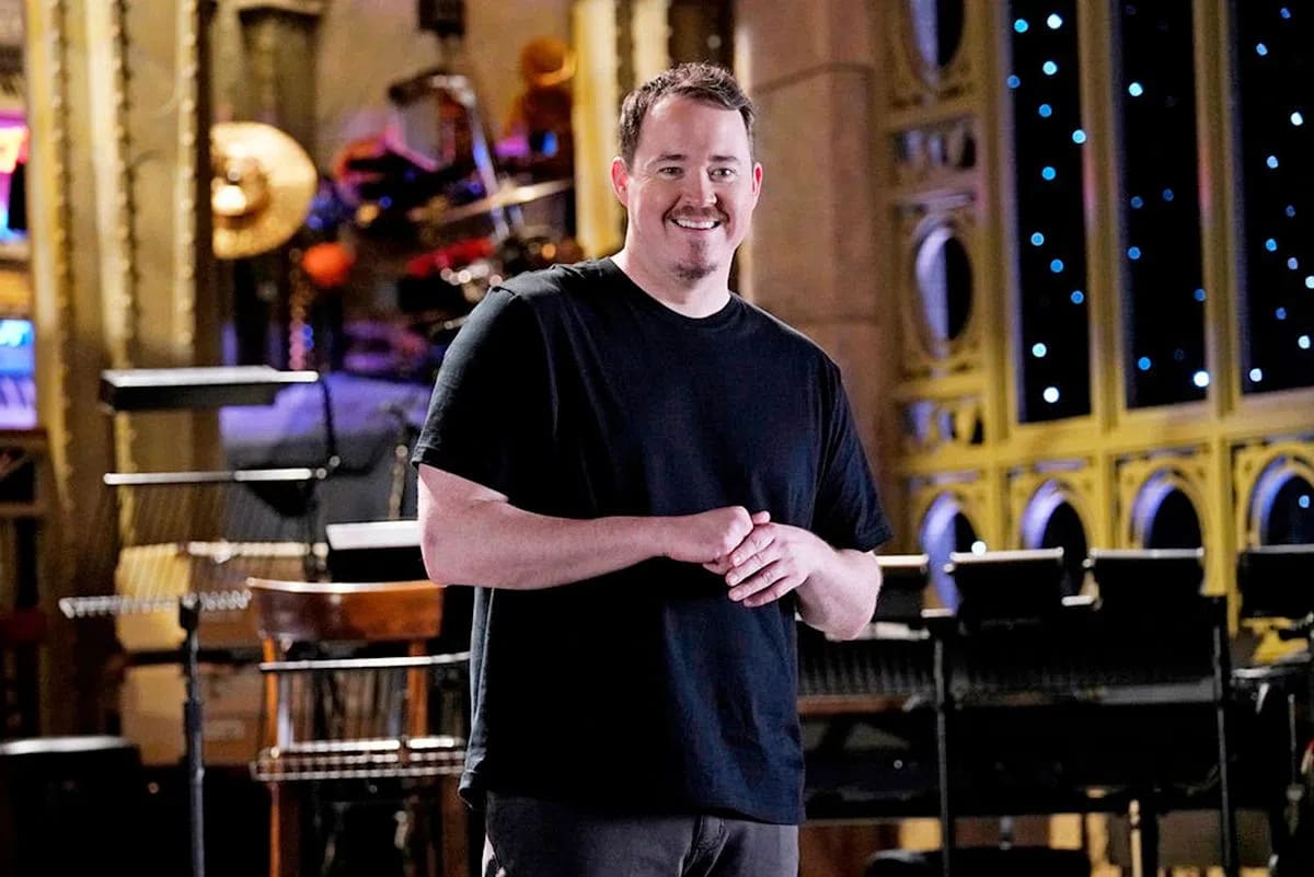 A man with short brown hair, a mustache, and a soul patch smiles widely while standing on a stage with his hands clasped together in front of him. He is wearing a plain black crewneck t-shirt and grey jeans. The stage behind him features musical instruments, microphone stands, an orchestral setup, and tall, gold-trimmed arched windows adorned with small blue lights that resemble a starry night sky.