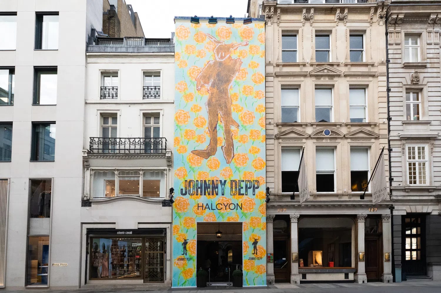 A tall, narrow building facade in an urban streetscape is completely covered by a vibrant, full-height art installation banner by Johnny Depp for his "Halcyon" exhibition. The colorful banner features a light blue background densely patterned with bright orange rose motifs, framing a central, stylized brown silhouette of a humanoid figure with long rabbit-like ears. Below the artwork, the text "JOHNNY DEPP" is printed in large, black, block lettering, directly above the word "HALCYON." A dark rectangular entryway is cut out at the base of the banner, leading into the gallery space. The decorated building is tightly flanked on both sides by traditional, multi-story European city architecture, including a white building with black iron balconies to its left and a classic stone building with rows of windows and white flags to its right, creating a stark contrast between classic street design and contemporary pop-art promotion.