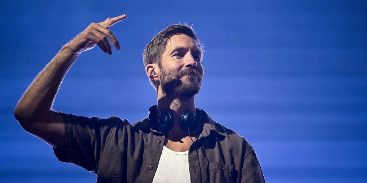 An outdoor musical performance features a man, captured from the chest up, with a short beard and mustache. He is wearing a dark, unbuttoned short-sleeved shirt over a white undershirt, with black headphones resting around his neck. His right arm is raised with his fingers pointing upward, and he has a subtle smile on his face. The backdrop is a solid vibrant blue with faint horizontal lines running across it, and a bright spotlight highlights his right side.