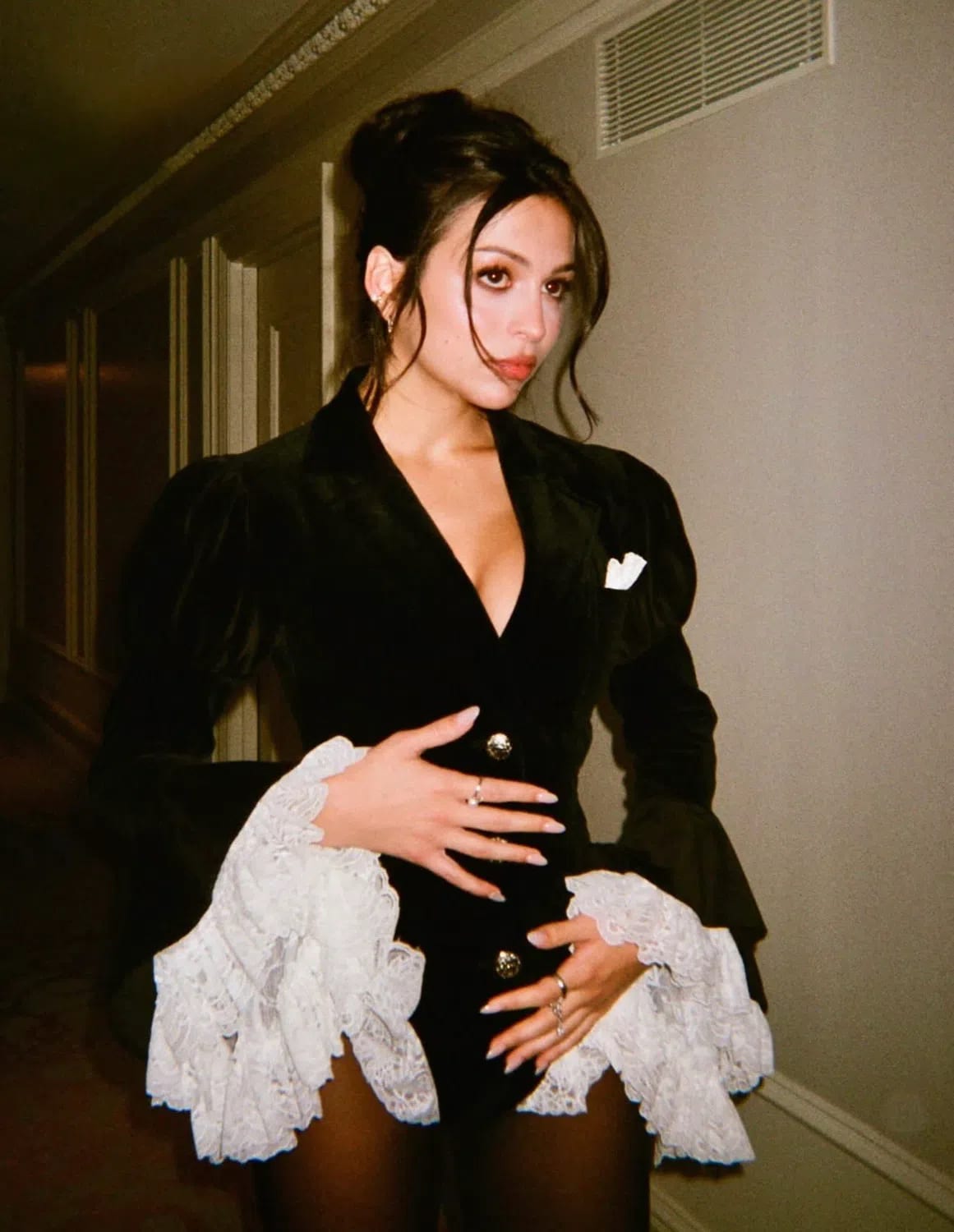 A flash-photographed, medium shot of American actress Josie Totah looking slightly to her right while posing indoors against a neutral, beige wall. She has dark hair styled in an elegant updo with loose, wavy face-framing tendrils. She wears a structured black velvet blazer or mini dress featuring a deep V-neckline, prominent puffed shoulders, and ornate gold buttons fastening the front. A small white pocket square accents the left side of the chest. The long sleeves flare dramatically at the wrists into tiered, voluminous white lace ruffles. She rests her manicured hands with long, light-colored nails and multiple metallic rings over her torso, over thin black sheer tights. A ventilation grate is visible on the upper right wall in the softly lit background.