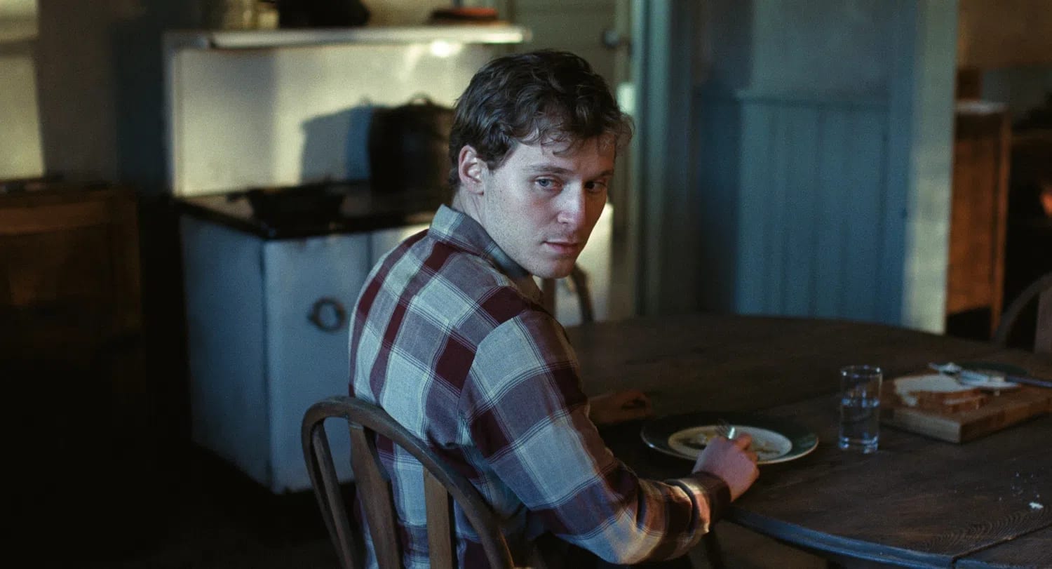 A medium shot captures actor Fred Hechinger as Mike sitting at a rustic wooden dinner table inside a dimly lit cabin kitchen, glancing over his shoulder with an intense, wary expression. He wears a brown, beige, and white plaid flannel shirt and holds a fork in his hand over a ceramic plate, while a half-empty glass of water sits next to a wooden cutting board with sliced bread nearby. The background is softly blurred, revealing a vintage white kitchen cabinet unit and a dark stove cast in shadows, emphasizing the somber and rustic 1950s period setting.