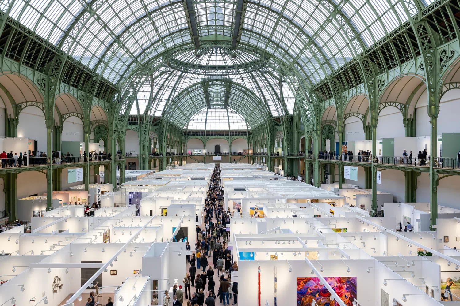A high-angle, wide-shot photograph captures the vast main exhibition floor of the Art Basel Paris art fair organized inside the sprawling nave of the historic Grand Palais in Paris, France. The massive space is covered by an ornate, vaulted glass and green-painted iron dome roof structure that floods the interior with natural light. Dozens of temporary, white-walled exhibition booths are arranged in a dense, geometric grid across the ground floor, creating corridors filled with visitors browsing contemporary artwork and sculptures. A long, straight central aisle cuts through the middle of the floor layout, packed with people walking between the various gallery displays. Surrounding the main floor, elevated perimeter balconies with green iron railings wrap around the structure, also populated with visitors overlooking the event. Large informational banners hang vertically from the upper railings on either side, while modern artwork, including vibrant abstract paintings and multi-colored wall hangings, is visible inside several open-fronted booths below.