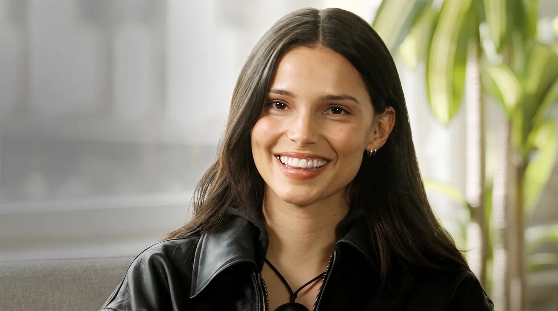 An elegant medium shot captures Inde Navarrette smiling warmly while looking directly forward against a softly blurred indoor backdrop. She has long, dark brown hair parted down the middle that drapes gracefully over her shoulders, framing her smooth complexion, defined cheekbones, and bright smile that reveals white teeth. Her ears are adorned with small gold hoop earrings, and she wears a collared black leather jacket over a matching top, styled with a simple dark cord necklace. The ambient lighting filters in from the side, illuminating her features and casting a gentle glow on the green leaves of a houseplant visible in the out-of-focus background.