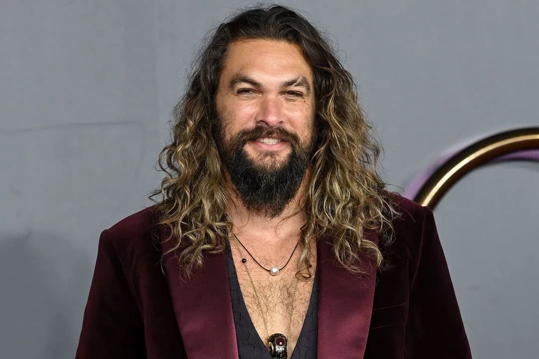 The image captures actor Jason Momoa from the chest up as he poses against a neutral gray background with a hint of a metallic gold structure behind him. He sports his signature long, wavy, sun-bleached brown hair that falls past his shoulders, paired with a full, thick beard and mustache. He is looking directly at the camera with a gentle, warm smile. Momoa wears a rich, deep burgundy velvet blazer featuring satin lapels over a low-cut dark top that reveals a portion of his chest. Layered around his neck are two distinct necklaces: a shorter black cord featuring a single white pearl or bead, and a longer golden chain holding a dark, ornate circular pendant.