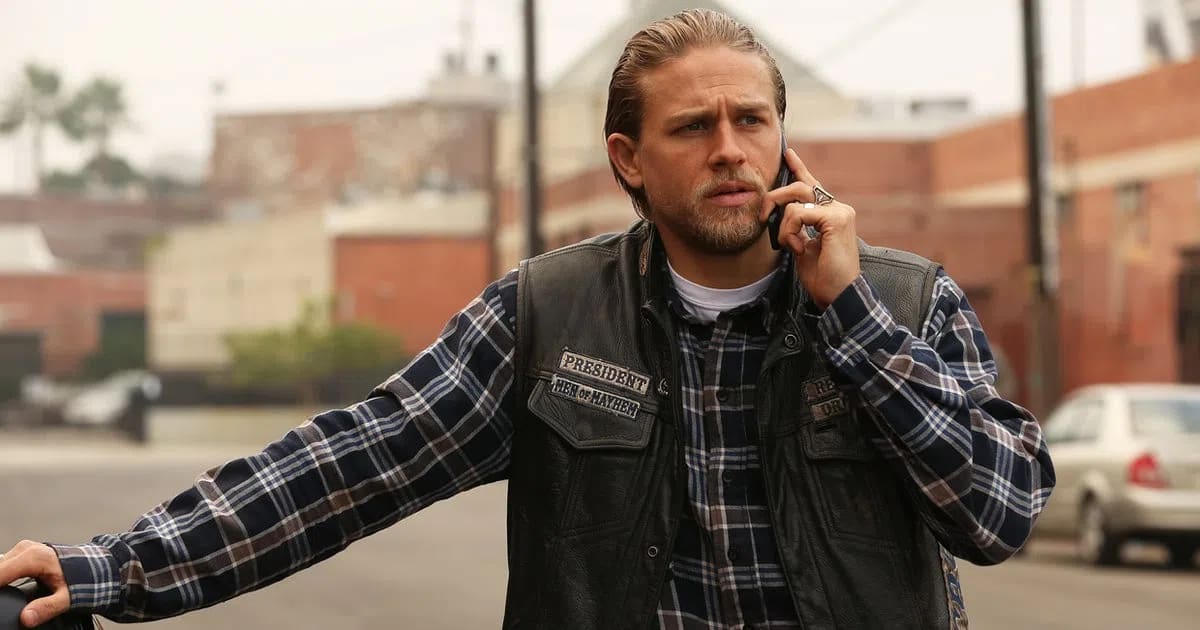 Jax Teller, the fierce president of the Sons of Anarchy Motorcycle Club, stands on a sun-drenched, gritty urban street while intensely focusing on a phone call. He sports his iconic biker look, donning a dark leather cuts vest layered over a blue, black, and white plaid flannel button-down shirt revealing a classic white crewneck undershirt beneath. His long blonde hair is slicked back neatly, complementing his rugged beard and mustache, while his left hand, adorned with prominent rings, holds the phone to his ear. The leather vest displays detailed, stitched club patches including "PRESIDENT" and "MEN OF MAYHEM" on the right breast, capturing his high-ranking status within the outlaw charter. In the soft-focus background, the hazy atmosphere of Charming features industrial buildings, sparse palm trees under an overcast sky, and a parked silver sedan, perfectly encapsulating the dramatic and tense mood of the television series.